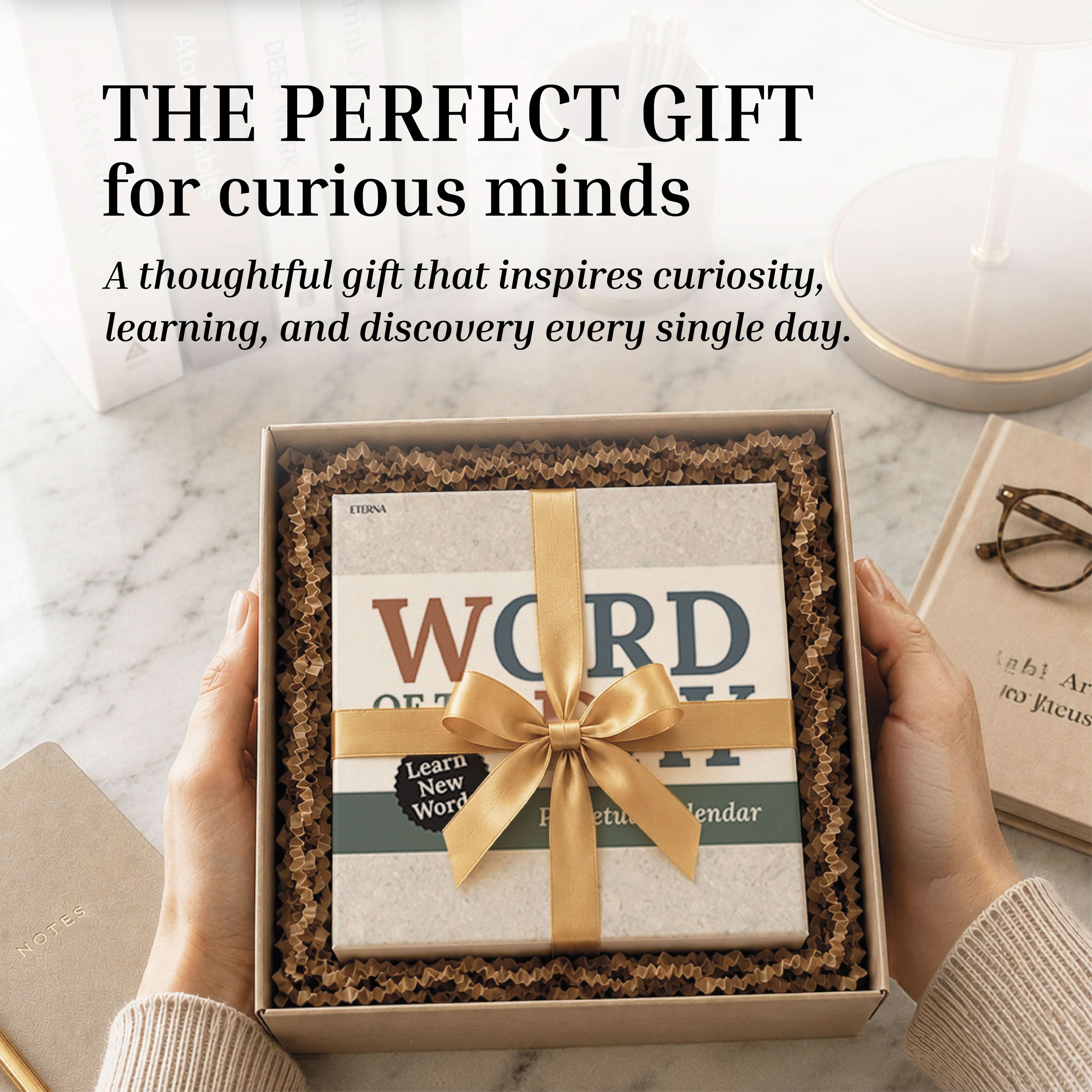 Hands hold a gift box containing the Perpetual Word of the Day Calendar by Eterna Goods—a thoughtful vocabulary builder—adorned with a gold ribbon, surrounded by books and a cup. The text highlights this calendar as an inspiring daily gift.