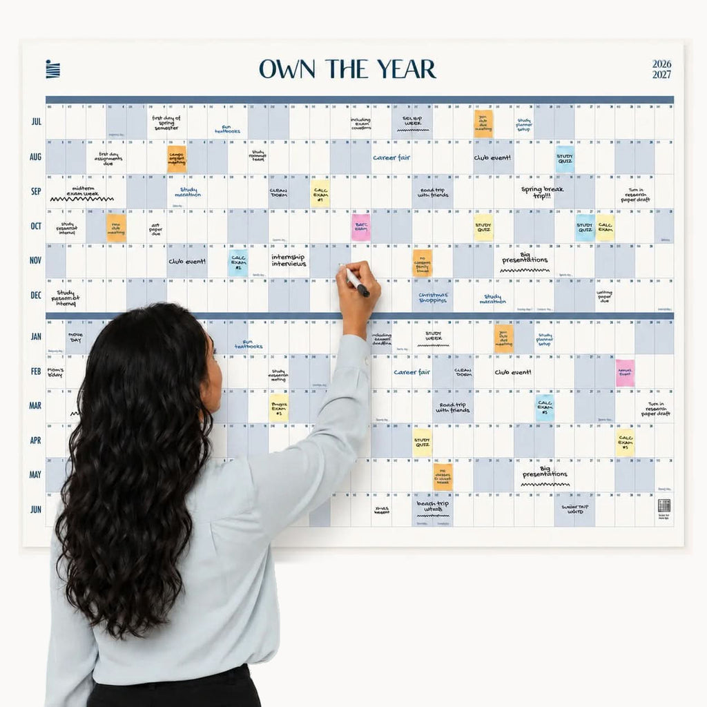 A woman with long hair writes on a large Eterna Goods 2026-2027 Midyear/Academic Own The Year Calendar, which is organized by month and filled with various colored notes and events.