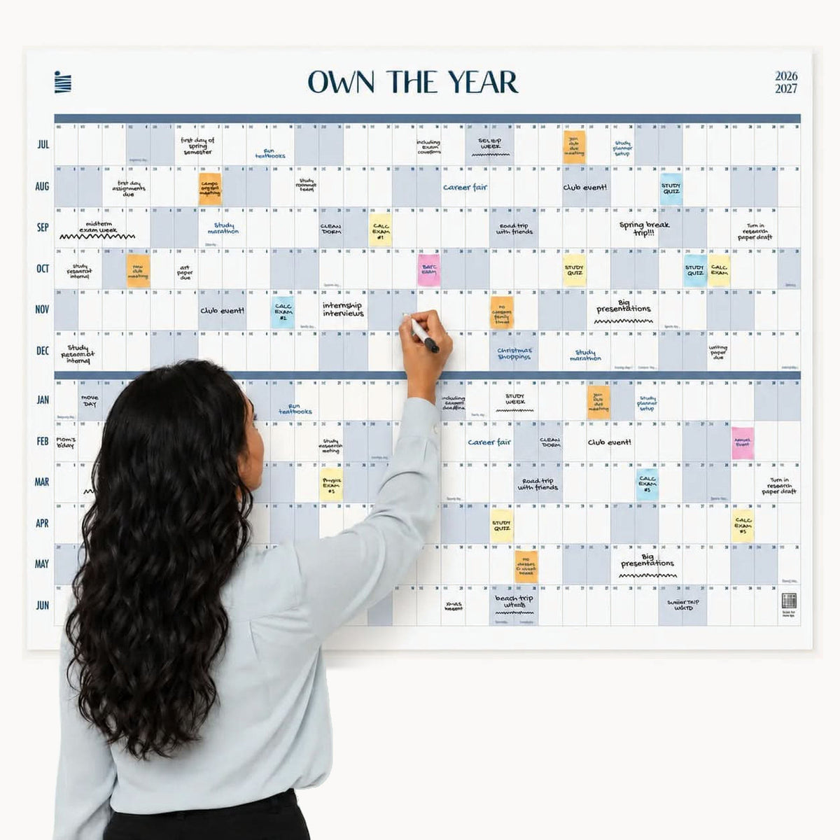 A woman with long hair writes on a large Eterna Goods 2026-2027 Midyear/Academic Own The Year Calendar, which is organized by month and filled with various colored notes and events.