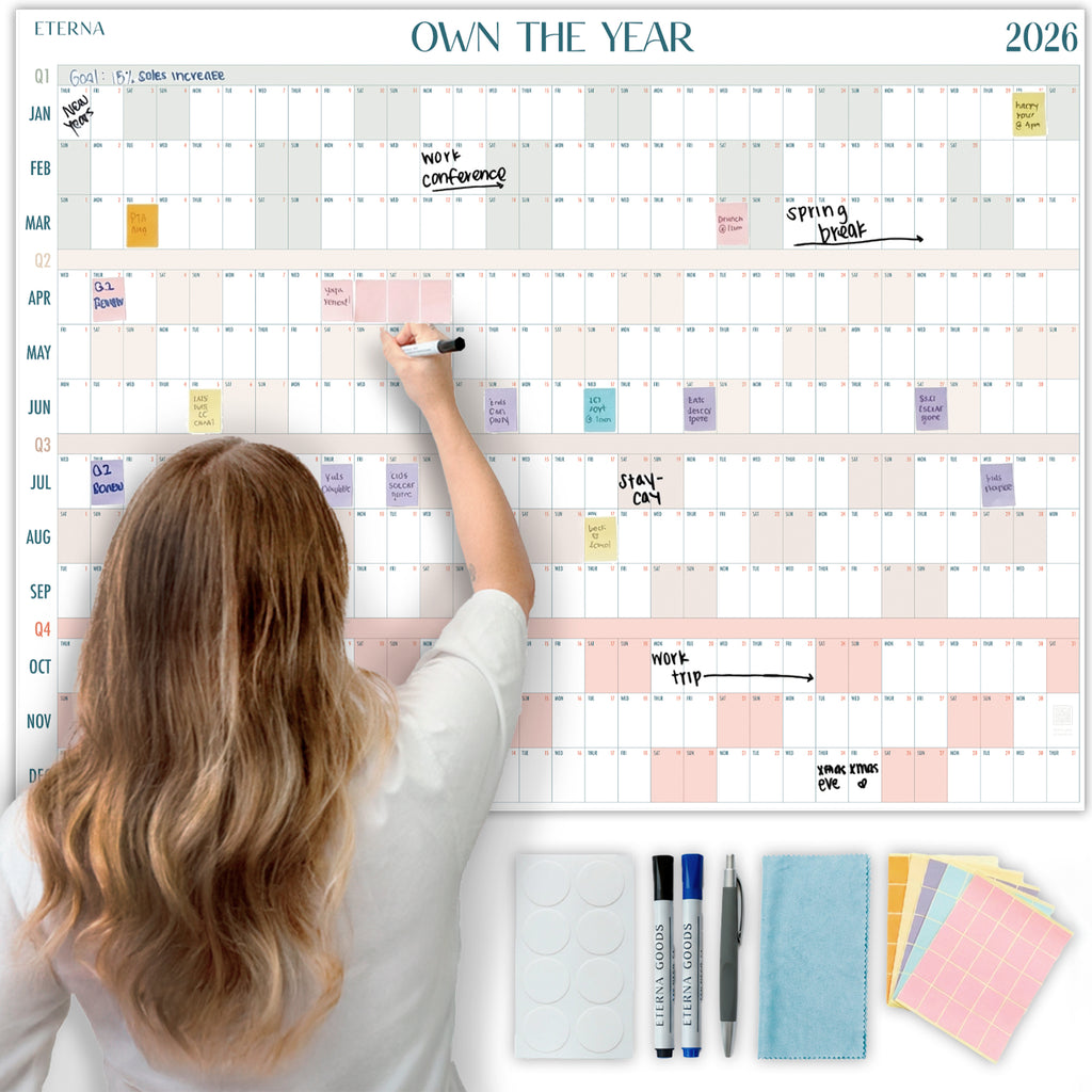 A woman writes on the Eterna Goods Own the Year 2026 GIANT Dry Erase Wall Calendar (4ft x 3ft), with events and notes displayed. Markers, erasers, stickers, and cloths are neatly arranged below for easy planning.