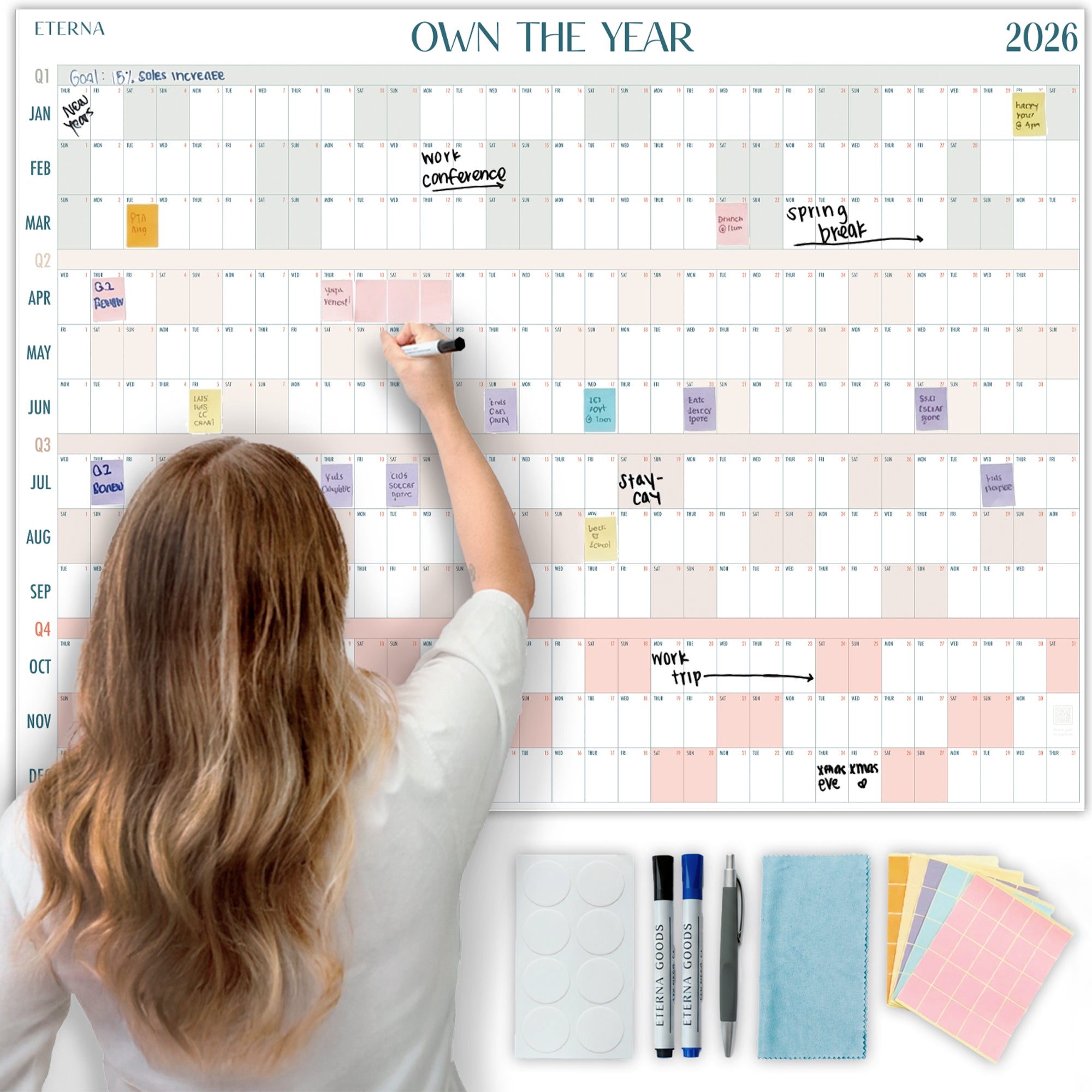 A woman writes on the Eterna Goods Own the Year 2026 GIANT Dry Erase Wall Calendar (4ft x 3ft), with events and notes displayed. Markers, erasers, stickers, and cloths are neatly arranged below for easy planning.