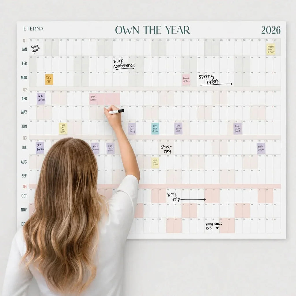 A woman writes with a marker on an Own The Year Calendar by Eterna Goods, a giant dry erase wall calendar mounted on the wall with various events and notes marked across multiple dates in 2026.