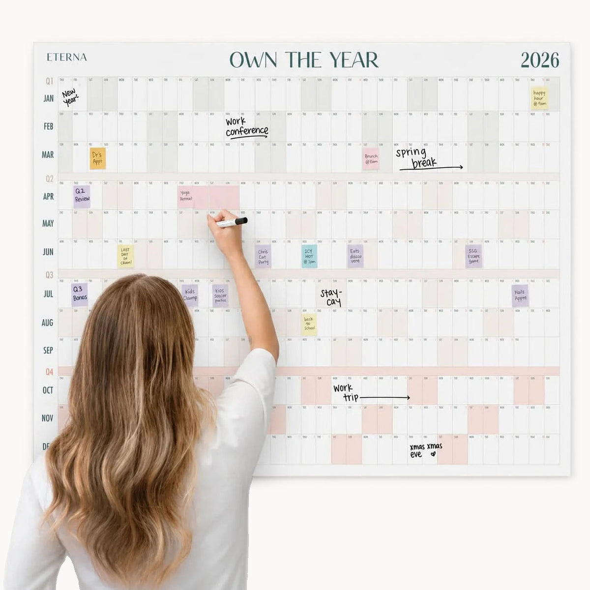 A woman writes on the Eterna Goods Own The Year Giant Dry Erase Calendar - Monster for 2026, which is organized by months and days and features various events already marked in colorful sticky notes and handwriting.