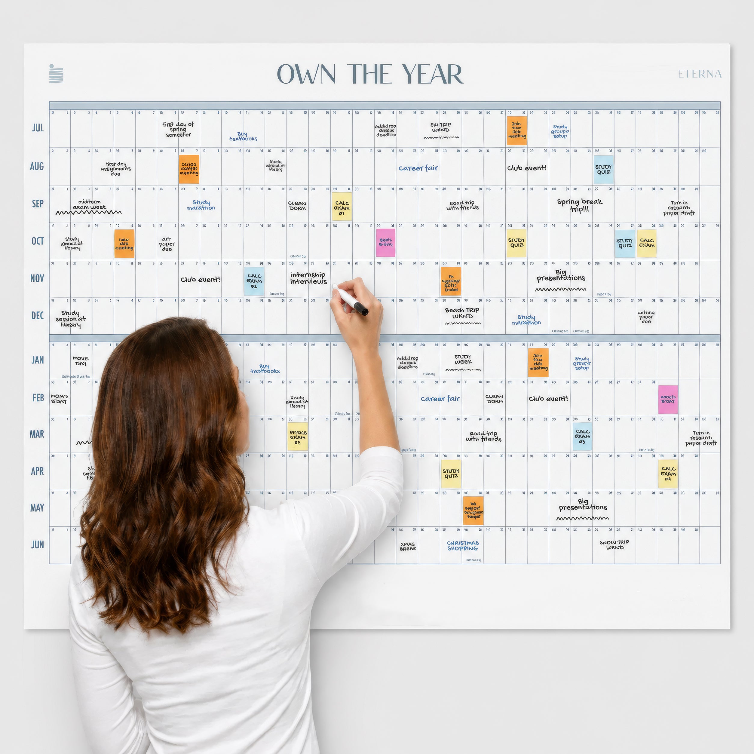 An individual writes on the Eterna Goods Undated Own The Year Calendar, a spacious wall calendar filled with colorful sticky notes and scheduled events, featuring clearly marked months and days along the sides and top, making it easy to update plans year-round with this versatile dry erase organizer.