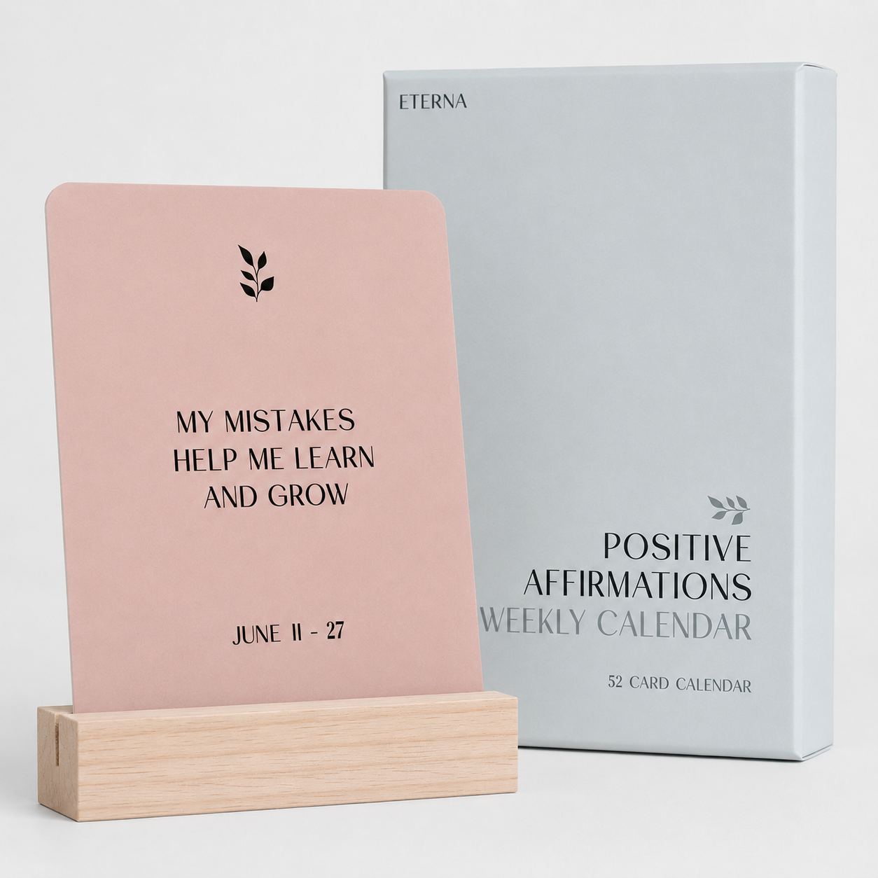 A pink Eterna Goods affirmation card says, "My mistakes help me learn and grow. June 11-27," on a wooden stand beside the Positive Affirmation Cards Weekly Calendar box—a thoughtful self-care gift to inspire daily mindfulness.