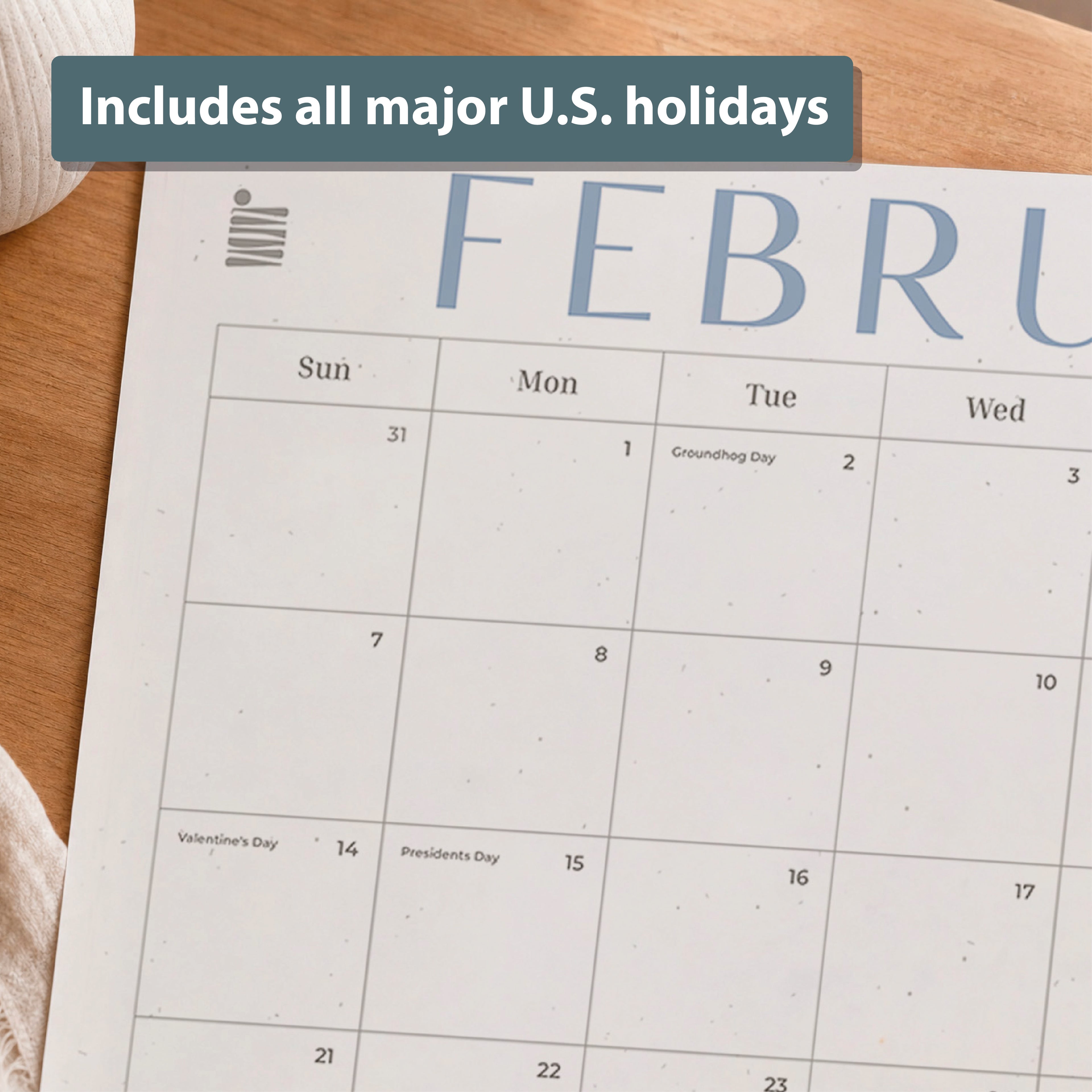 A close-up of the Eterna Goods 2026-2027 18-Month Flat Desk Calendar (22"x17") on a wooden desk, featuring a February page with marked U.S. holidays like Groundhog Day, Valentine's Day, and Presidents' Day and plenty of writing space.