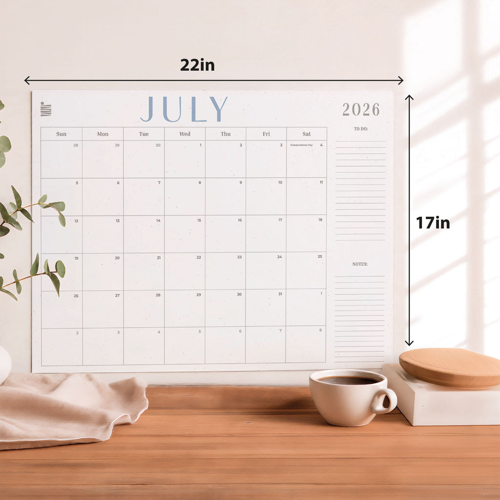 The Eterna Goods 2026-2027 18-Month Flat Desk Calendar (22"x17") with spacious writing areas is shown on a wooden desk alongside a coffee cup, plant, and fabric.