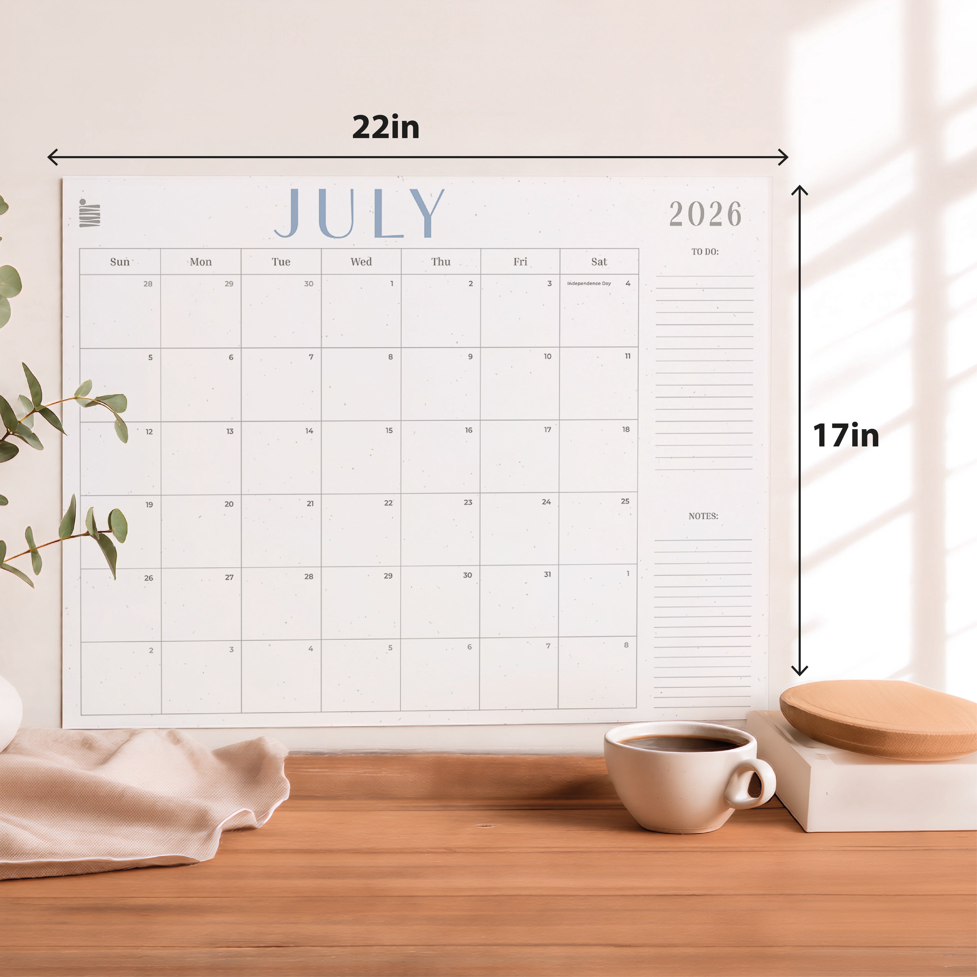 The Eterna Goods 2026-2027 18-Month Flat Desk Calendar (22"x17") with spacious writing areas is shown on a wooden desk alongside a coffee cup, plant, and fabric.