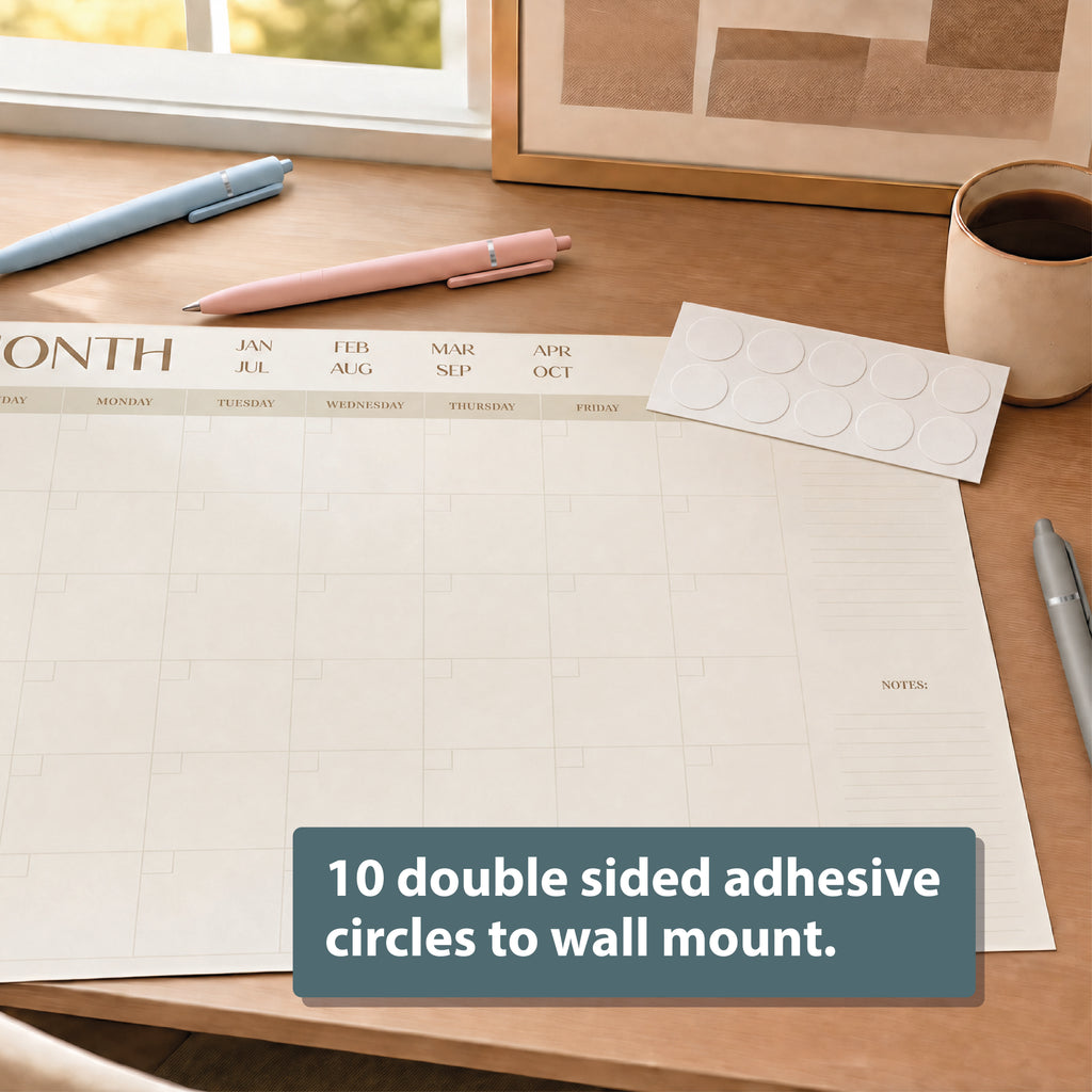 The Eterna Goods Undated 12-Month Flat Desk Calendar (22" x 17") sits on a desk with pens, a coffee cup, and a sheet of 10 double-sided adhesive circles for wall mounting.