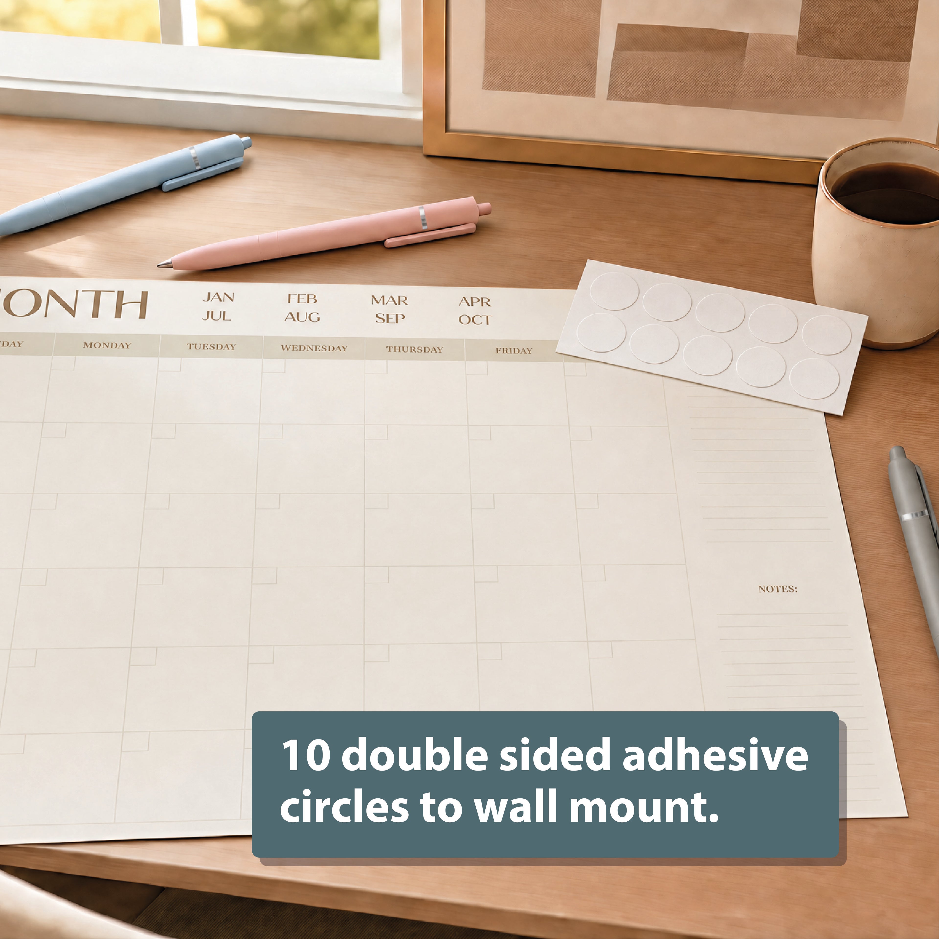 The Eterna Goods Undated 12-Month Flat Desk Calendar (22" x 17") sits on a desk with pens, a coffee cup, and a sheet of 10 double-sided adhesive circles for wall mounting.