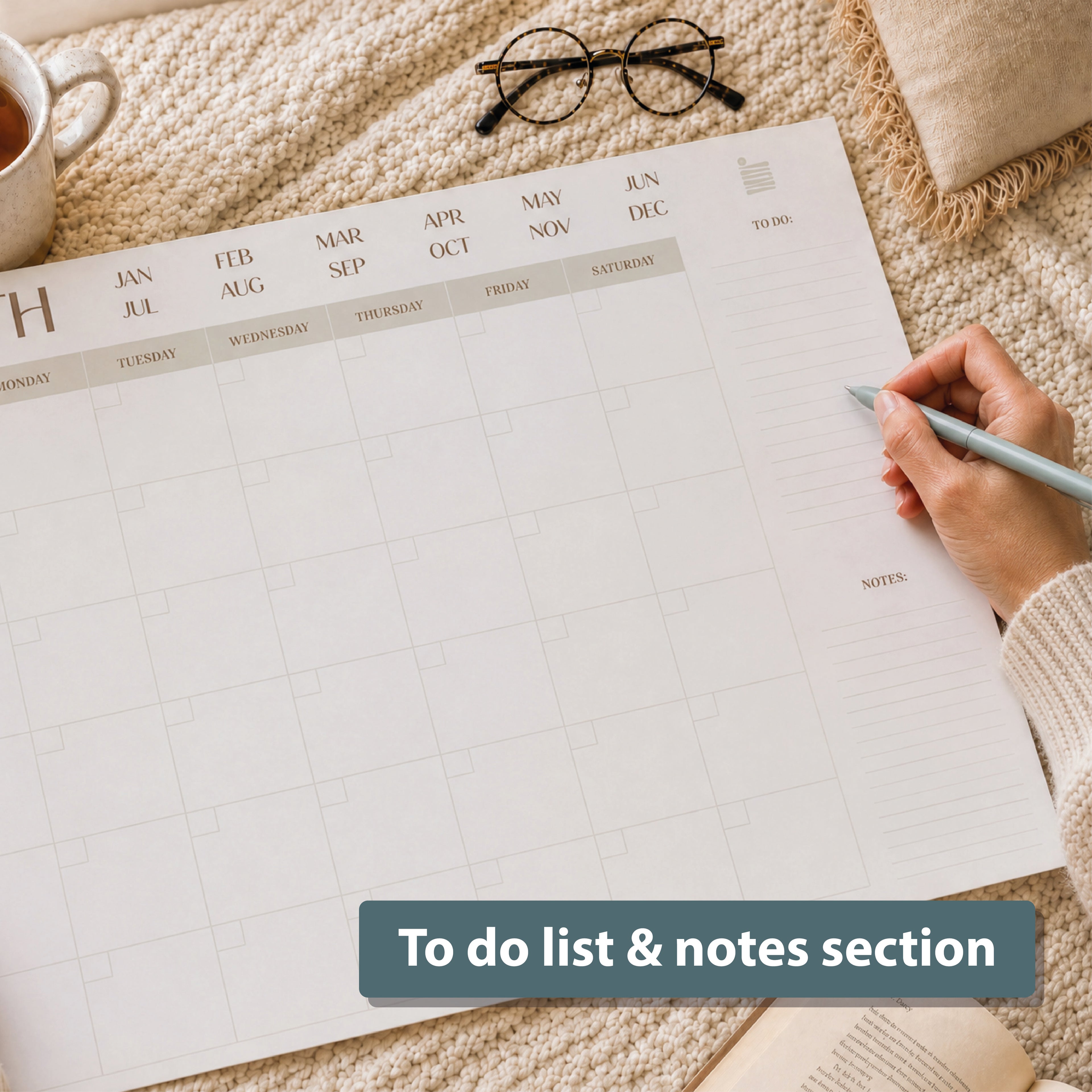 Writing on the Eterna Goods Undated 12-Month Flat Desk Calendar (22" x 17"), featuring spacious monthly pages for to-dos and notes. A cup of coffee, eyeglasses, and a cozy blanket add comfort nearby.
