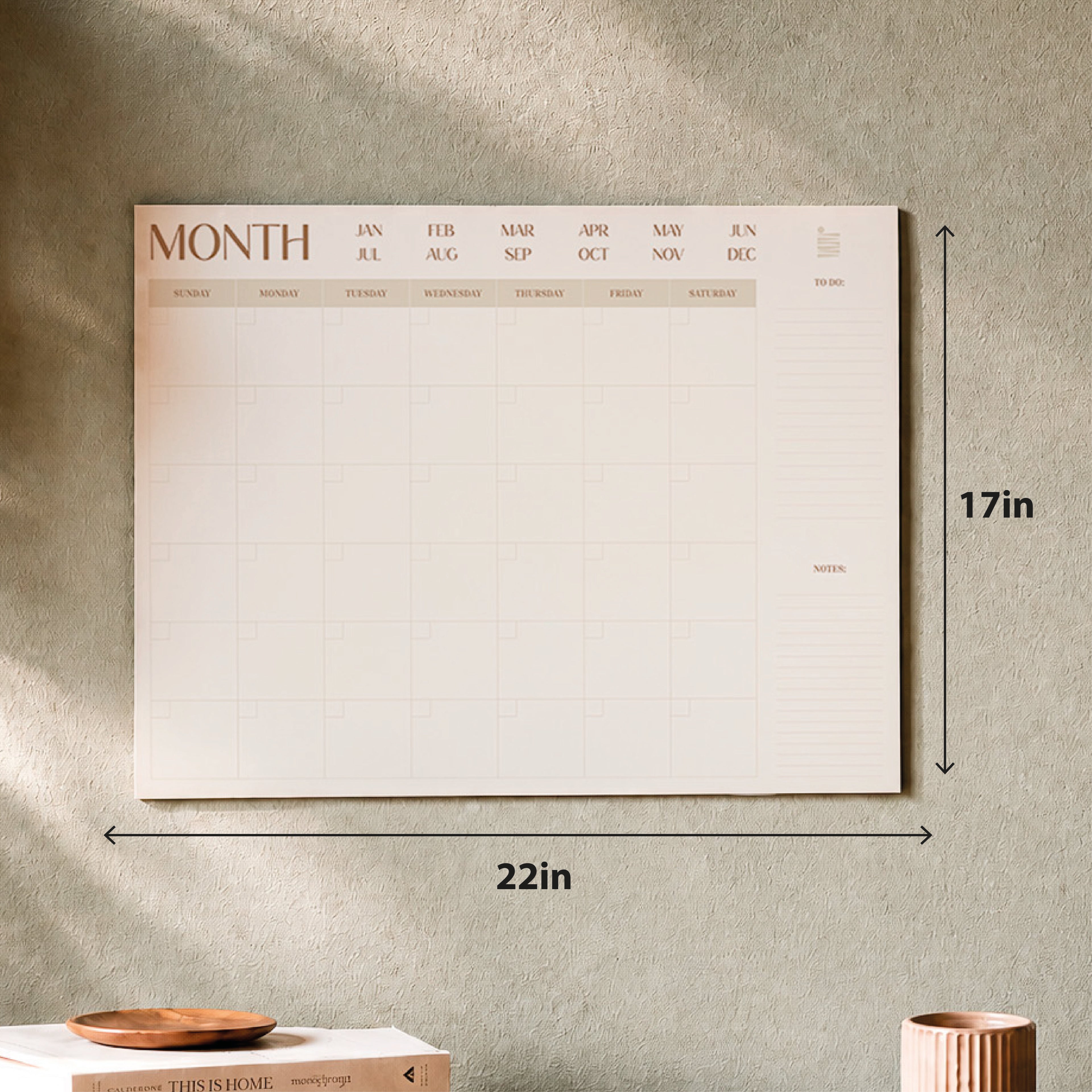 The Eterna Goods Undated 12-Month Flat Desk Calendar (22" x 17") is shown on a beige wall above a table with books and a ribbed ceramic container, offering ample space for notes and plans.