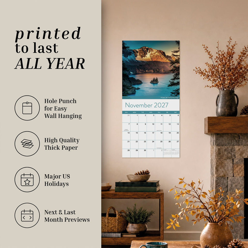 An Eterna Goods 2027 National Parks Wall Calendar hangs in the living room above a shelf displaying books, a plant, and autumn-inspired decor; ideal for outdoor enthusiasts, this calendar showcases breathtaking National Park photography with a mountain lake scene and product highlights.