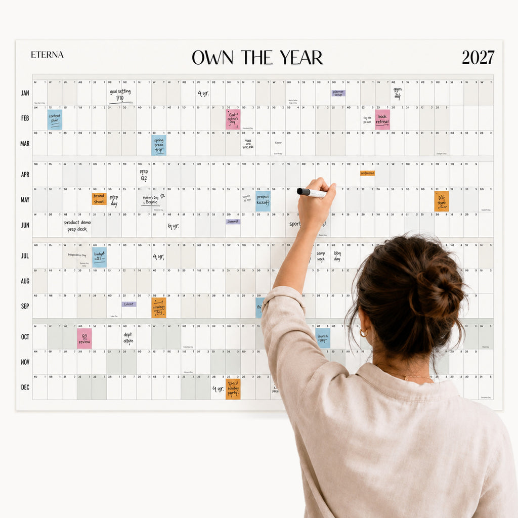 A person writes on the Eterna Goods Dated Own The Year Calendar for 2027, which is covered with colorful sticky notes and planning details.