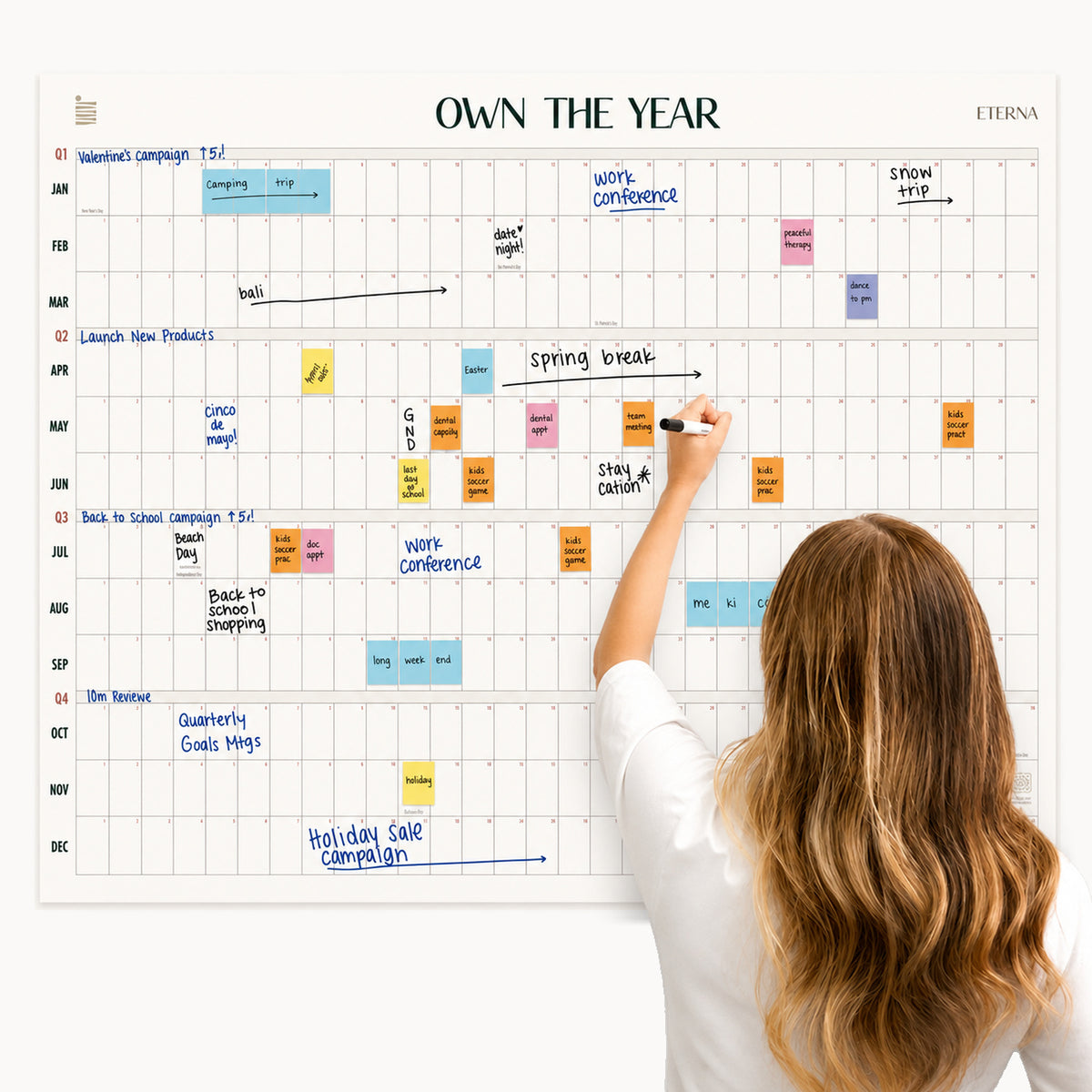 A woman writes on the Undated Own The Year Calendar by Eterna Goods, a wall-mounted yearly planner filled with events, tasks, and notes written in various colors. | variant:Original
