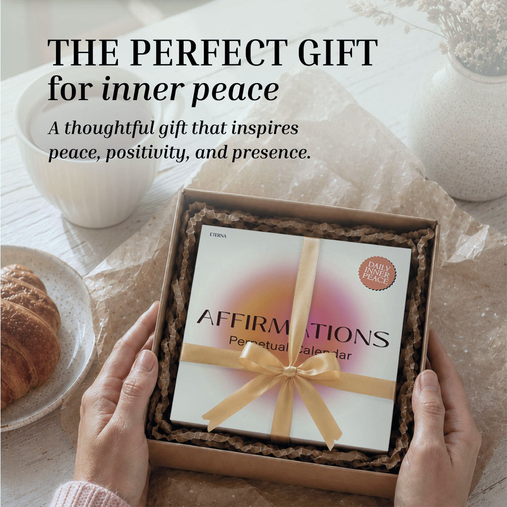 A person holds a gift box featuring the Eterna Goods Perpetual Affirmations Calendar, beautifully tied with a ribbon to offer daily inspiration, while a croissant and a vase sit on the table nearby.