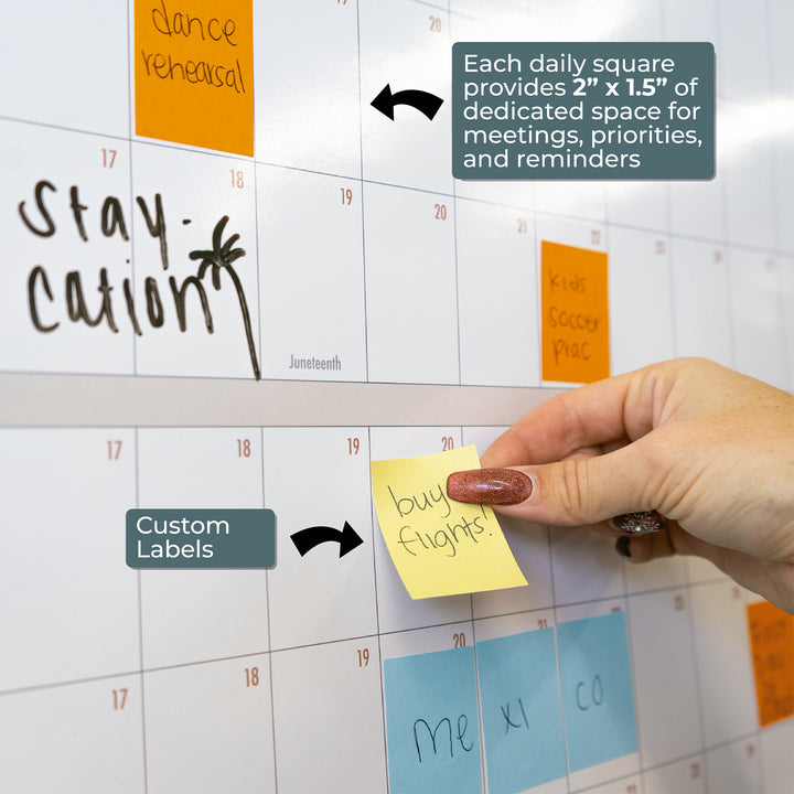 A hand places a "buy flights" sticky note on the Eterna Goods Own the Year 2026 GIANT Dry Erase Wall Calendar, a 4ft x 3ft yearly planner with custom labels and 2" x 1.5" daily squares for notes and reminders.