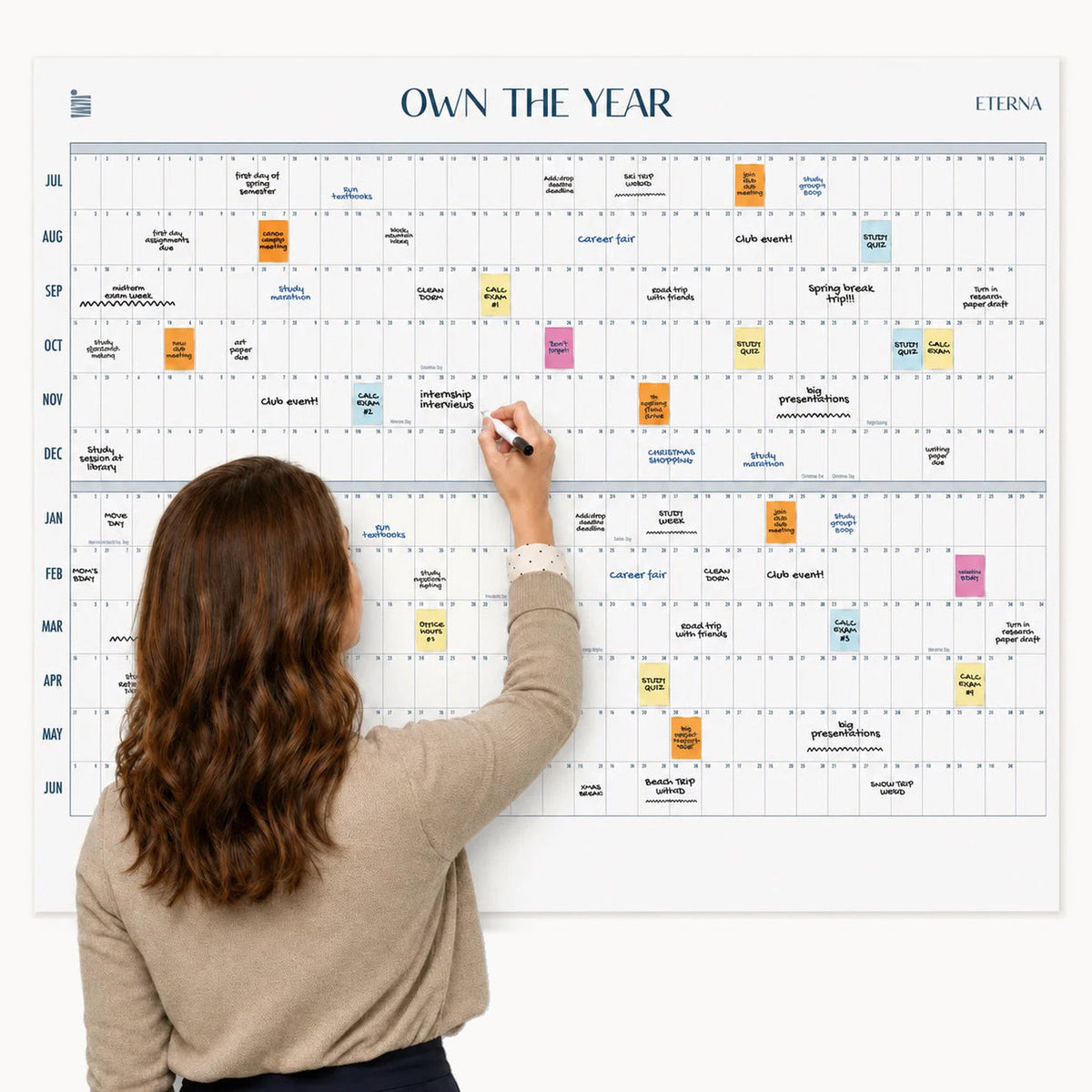 A woman writes on an Eterna Goods Undated Midyear/Academic Own The Year Calendar, a giant dry erase wall calendar that displays a year’s schedule with colorful event notes for each month.